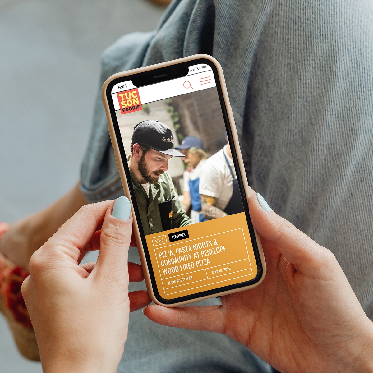 Tucson Foodie website homepage displayed on an iphone that is being held face forward by a person who is sat down cross legged.